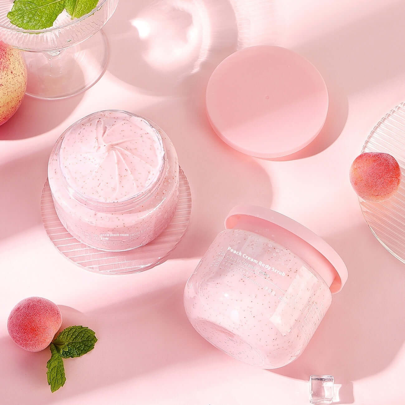 Peach Cream Body Scrub in a stylish pink jar on a pastel background, featuring fresh peaches and mint leaves.